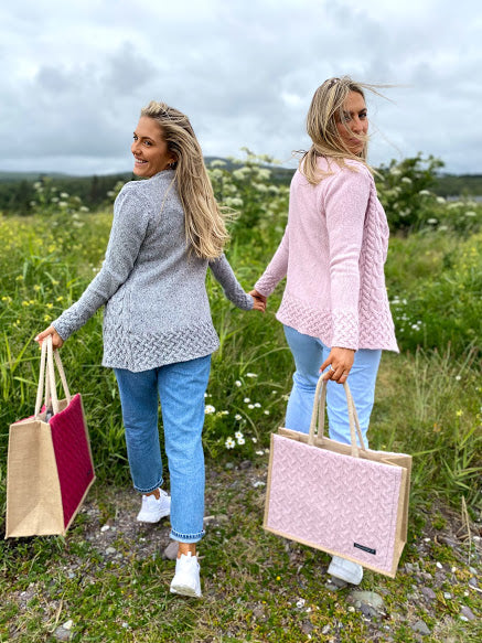 2 girls on Templenoe Beach Kenmare holding hands and looking back smiling ,one wearing a grey cardigan and carrying a berry wool bag and the other wearing a pink cardigan and carrying a pink bag Stepping from the stony beach into grass with wild daisies .