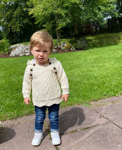 baby standing in the garden in jeans and white runners and an aran hoodie with buttons.colour natural with buttons from the neck to underarms on each side .